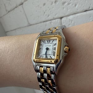 May include: A Cartier Panth&egrave;re watch with a silver and gold band. The watch face is square with Roman numerals and gold accents. The watch is on a wrist.