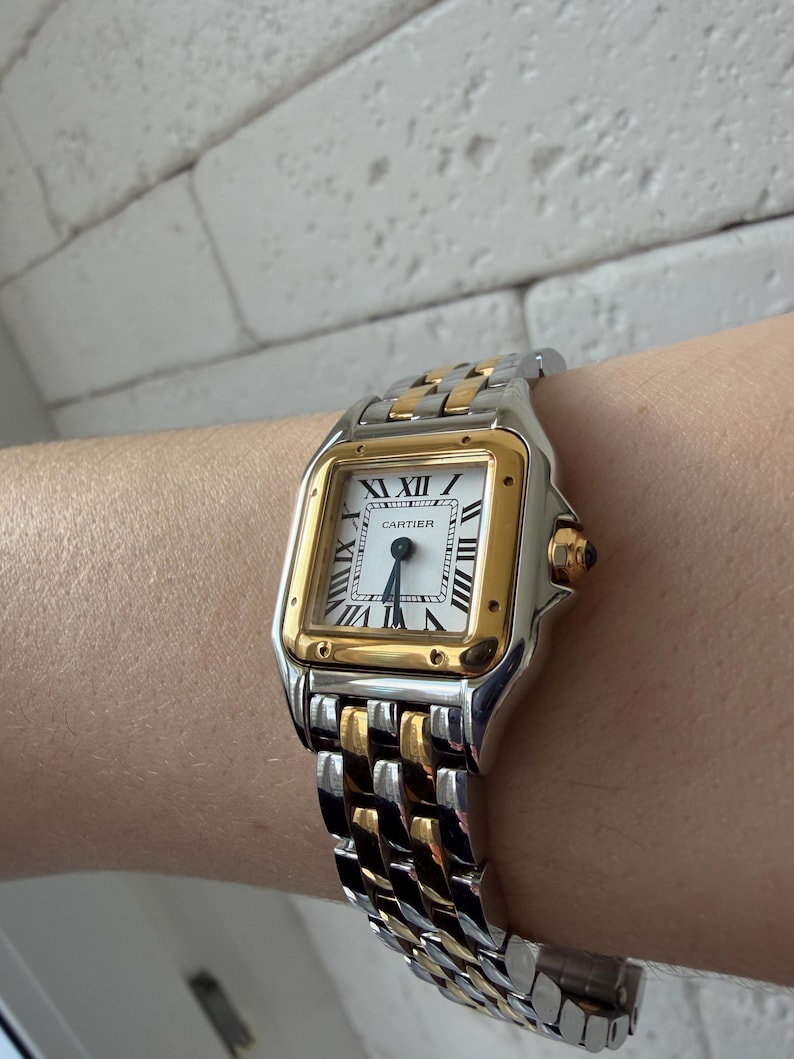 May include: A Cartier Panth&egrave;re watch with a silver and gold band. The watch face is square with Roman numerals and gold accents. The watch is on a wrist.