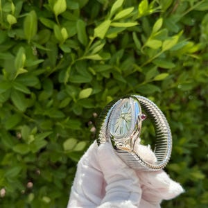 May include: A silver wristwatch with a snake-like design. The watch features a white face with silver hands and hour markers, and a small red gemstone. The band is a flexible, textured metal. The watch is held against a backdrop of green foliage.