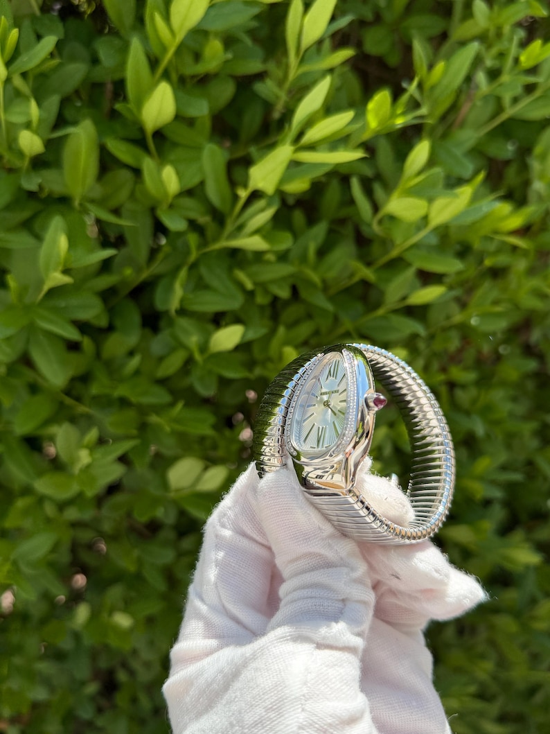 May include: A silver wristwatch with a snake-like design. The watch features a white face with silver hands and hour markers, and a small red gemstone. The band is a flexible, textured metal. The watch is held against a backdrop of green foliage.