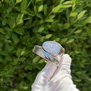 May include: A silver wristwatch with a unique oval face and a diamond-studded bezel. The watch features a light blue dial with silver hands and hour markers. The band is a flexible, ribbed design. The watch is displayed against a backdrop of green foliage.