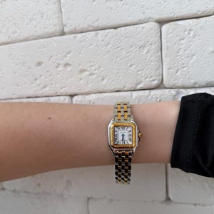 May include: A two-tone gold and silver wristwatch with a square face and Roman numeral hour markers. The watch is on a silver and gold link band. The watch is on a wrist.
