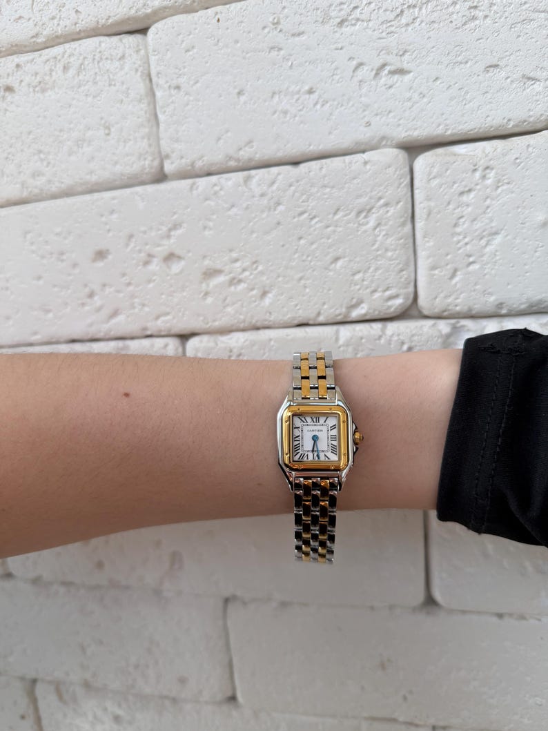 May include: A two-tone gold and silver wristwatch with a square face and Roman numeral hour markers. The watch is on a silver and gold link band. The watch is on a wrist.