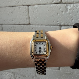 May include: A two-tone Cartier Panth&egrave;re watch with a gold square bezel and a silver dial featuring black Roman numerals. The watch has a gold crown and a stainless steel and gold link bracelet.