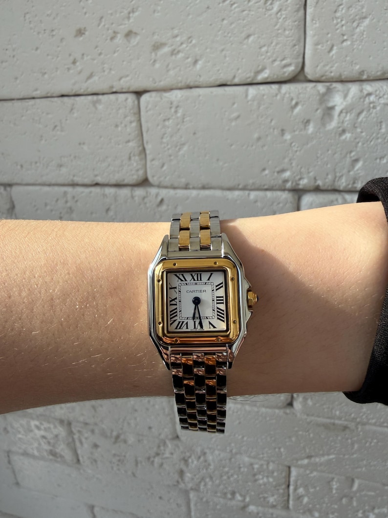 May include: A two-tone Cartier Panth&egrave;re watch with a gold square bezel and a silver dial featuring black Roman numerals. The watch has a gold crown and a stainless steel and gold link bracelet.