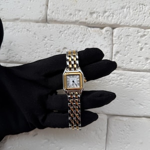 May include: A gold and silver wristwatch with a white face and Roman numeral hour markers. The watch has a rectangular face with a gold bezel and a two-tone metal band. The watch is held against a black background.