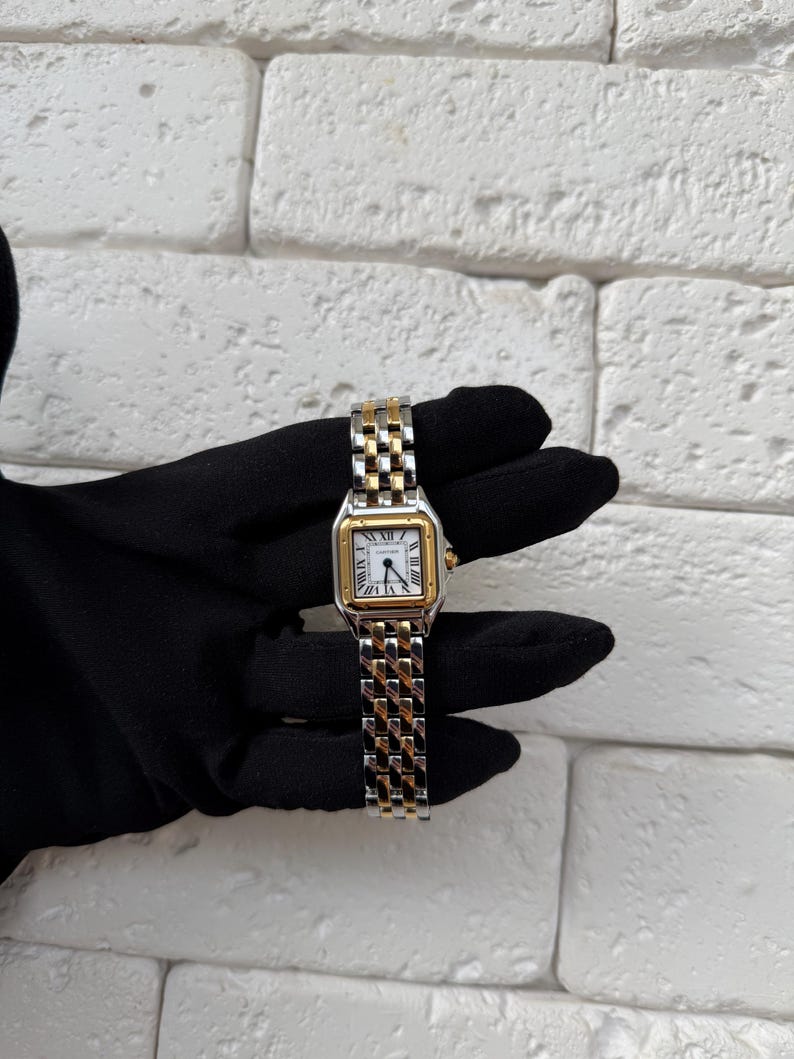 May include: A gold and silver wristwatch with a white face and Roman numeral hour markers. The watch has a rectangular face with a gold bezel and a two-tone metal band. The watch is held against a black background.