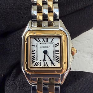 May include: A luxury wristwatch with a silver and gold band and a square white face. The watch face features black Roman numerals and the word "CARTIER" in black. The watch is marked "SWISS MADE".