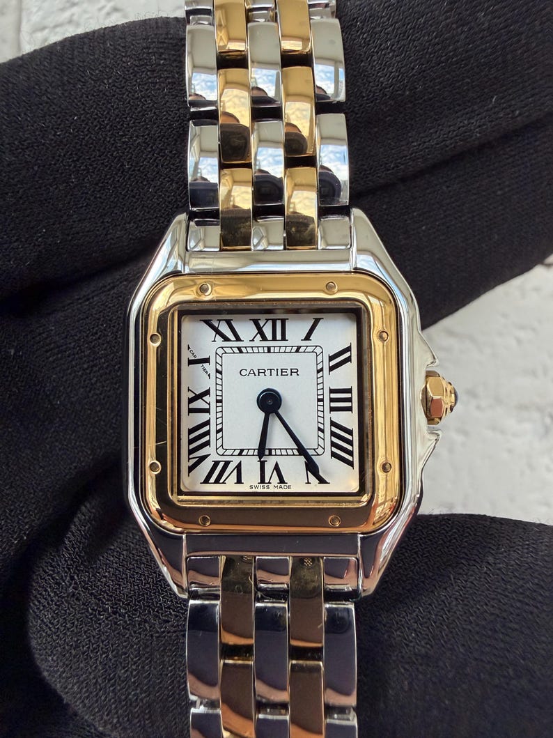 May include: A luxury wristwatch with a silver and gold band and a square white face. The watch face features black Roman numerals and the word "CARTIER" in black. The watch is marked "SWISS MADE".
