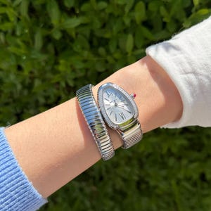May include: A silver wristwatch with a snake-like design, featuring a white dial and a bezel adorned with small, sparkling stones. The watch is paired with a matching silver bracelet. The watch and bracelet are on a wrist, with a green leafy background.