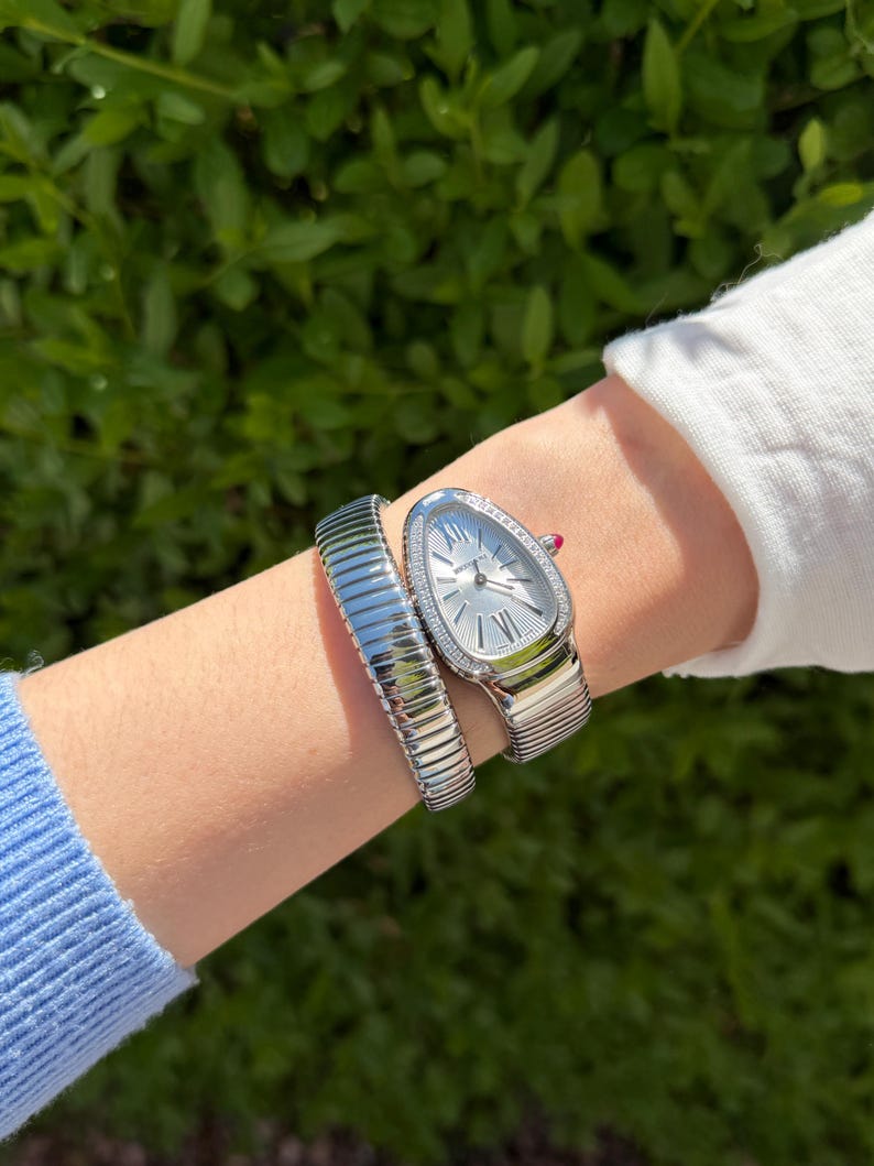 May include: A silver wristwatch with a snake-like design, featuring a white dial and a bezel adorned with small, sparkling stones. The watch is paired with a matching silver bracelet. The watch and bracelet are on a wrist, with a green leafy background.