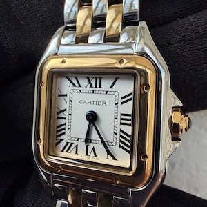 May include: A luxury wristwatch with a square white face, black Roman numerals, and black hands. The watch has a gold bezel and a two-tone gold and silver metal band. The watch face reads "CARTIER" and "SWISS MADE".