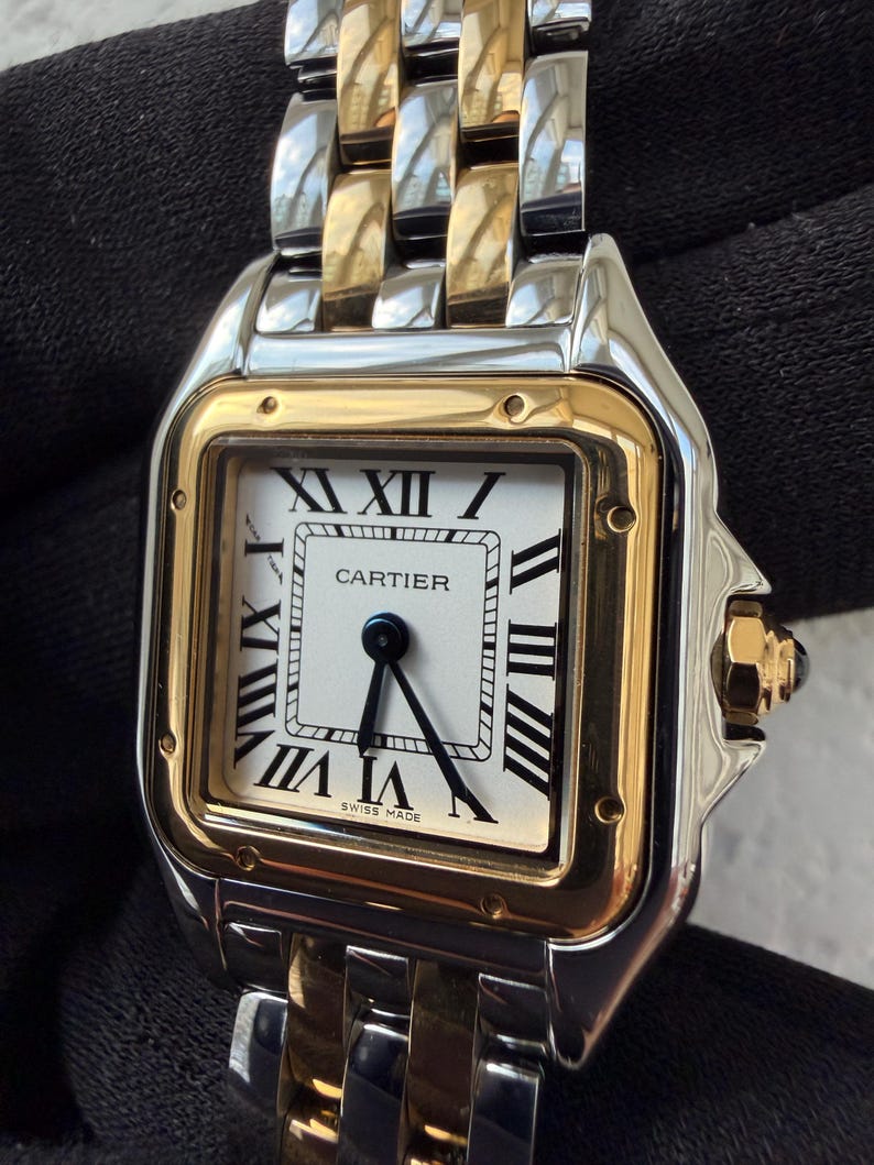 May include: A luxury wristwatch with a square white face, black Roman numerals, and black hands. The watch has a gold bezel and a two-tone gold and silver metal band. The watch face reads "CARTIER" and "SWISS MADE".