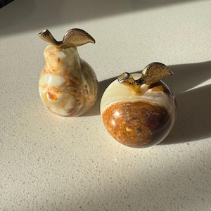 Könnte beinhalten: Zwei dekorative Obstskulpturen aus Stein. Eine ist birnenförmig, die andere apfelförmig. Beide haben ein goldfarbenes Blattdetail oben. Die Obstskulpturen sind aus einem cremefarbenen, braunen und beigefarbenen Stein gefertigt.