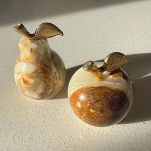 May include: Two decorative fruit figurines made of polished stone with natural brown, cream, and white veining. One figurine is shaped like a pear and the other like an apple, both topped with a textured, metallic gold-coloured stem and leaf.