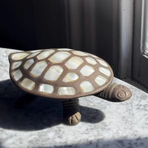 Könnte beinhalten: Eine Schildkrötenfigur mit einem dunkelbraunen Metallkörper und einem Panzer, der mit schillernden weißen Perlmuttplättchen eingelegt ist. Die Schildkröte hat einen strukturierten Kopf mit kreisförmigen Augendetails und vier Beine sowie ein geometrisches Muster.