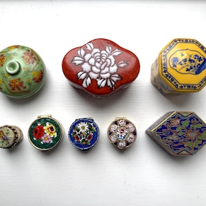 Puede incluir: Colección de joyeros decorativos de varias formas y tamaños. Incluye una caja redonda verde con diseño floral, una caja roja con una flor blanca y una caja octogonal amarilla con patrones florales azules. Las cajas más pequeñas presentan diseños intrincados.