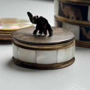 May include: A small, round trinket box with a lid featuring a bronze elephant figurine. The box's body is adorned with rectangular mother-of-pearl tiles, and it has a bronze-colored rim. Other similar boxes are in the background.
