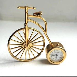 May include: A gold-colored miniature bicycle clock with a large front wheel and a small rear wheel. The clock face is white with black Roman numerals and hands. The bicycle has a seat and handlebars.