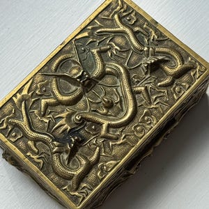 May include: Rectangular brass box with intricate relief carvings of dragons and foliage. The box has a hinged lid and a detailed, textured surface. The color is a warm, antique gold.