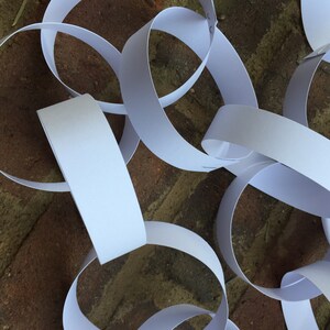 May include: A close-up of a white paper chain, made of multiple linked paper rings, lying on a brown brick surface.