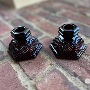 May include: Two dark red glass candle holders with a hexagonal base and intricate designs. The holders are sitting on a brick surface.