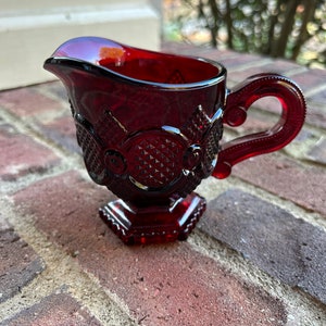 May include: A small, red glass creamer with a decorative pattern and a handle. The creamer has a footed base.