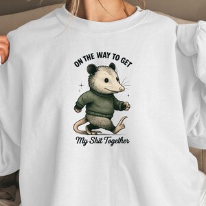 Pode incluir: Camisola branca com um desenho de um gambá de desenho animado usando um suéter verde, andando com uma expressão determinada. O texto acima diz "ON THE WAY TO GET", e abaixo, "My Shit Together".