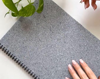 Può includere: Un quaderno a spirale con una copertina grigia testurizzata e maculata. Il quaderno presenta una rilegatura in filo metallico nero sul lato sinistro. La copertina ha un aspetto screziato con varie tonalità di grigio e una superficie granulosa.
