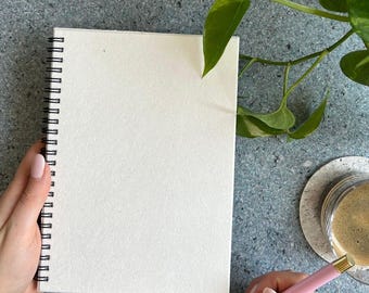 Può includere: Un taccuino a spirale con una copertina bianca semplice e una superficie ruvida. Il taccuino è tenuto insieme a una penna con il corpo rosa chiaro e l'impugnatura verde. Il taccuino è rettangolare con una rilegatura a spirale nera laterale.