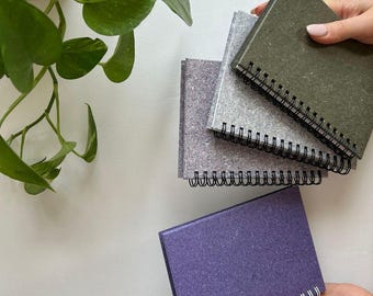 Può includere: Un set di quattro piccoli taccuini rilegati a spirale con copertine testurizzate nei toni del verde oliva, grigio chiaro, grigio scuro e viola. I taccuini presentano una rilegatura a spirale nera per i tre superiori e bianca per quello viola.