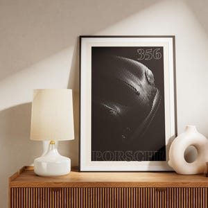 Könnte beinhalten: Gerahmter schwarz-weißer Porsche 356 Kunstdruck. Der Druck zeigt ein stilisiertes Bild der Fahrzeugfront mit der Nummer "356" oben und "PORSCHE" unten. Eine weiße Lampe und Vase stehen auf einem Holzschrank.