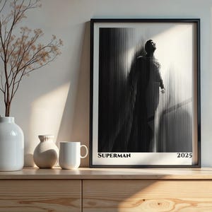 Könnte beinhalten: Schwarz gerahmter Kunstdruck mit einem stilisierten Superman-Bild. Das Kunstwerk zeigt eine Figur in einem schwarz-weißen Streifenmuster. Das Wort "SUPERMAN" und das Jahr "2025" sind unten aufgedruckt.