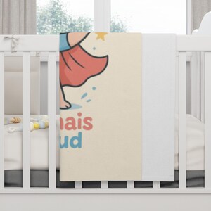 May include: A baby blanket with a cartoon illustration of a figure in a red skirt and blue top. The blanket has the words "mais" and "ud" in red and blue. The blanket is cream-colored with a white border.