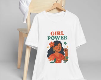 Puede incluir: Camiseta blanca con las palabras "GIRL POWER" en rojo y verde. La camiseta presenta un gráfico de una mujer con cabello oscuro, flores y un puño en alto. Una gorra blanca descansa sobre el respaldo de una silla de madera.