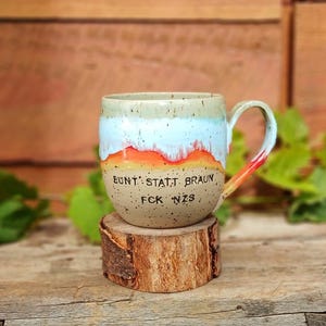 Könnte beinhalten: Eine handgefertigte Keramiktasse mit farbenfrohem Design. Die Tasse hat einen hellgrünen oberen Teil, eine weiße und orangefarbene Mitte und eine gesprenkelte beige Basis. Der Griff ist rot und orange. Die Tasse steht auf einer kleinen Holzscheibe. Der Text auf der Tasse lautet "BUNT STATT BRAUN FCK NZS."