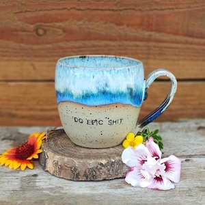 Könnte beinhalten: Handgefertigte Keramiktasse mit blau-weißer Glasur und dem Schriftzug "DO EPIC SHIT". Die Tasse steht auf einer Holzscheibe mit Blumen. Der Henkel hat blaue Akzente.