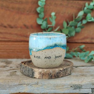 Könnte beinhalten: Eine Keramiktasse mit hellblauem und beigefarbenem, gesprenkeltem Design. Die Oberseite der Tasse hat eine hellblaue Glasur, die an der Seite heruntertropft. Die Worte "RISE. AND. SHINE" sind auf die Vorderseite der Tasse gedruckt.
