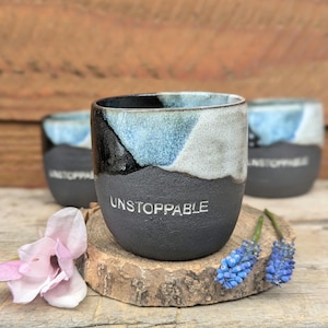 Könnte beinhalten: Drei Keramikbecher mit schwarzem Boden, einem hellblauen und weißen oberen Bereich und dem Wort "UNSTOPPABLE" auf der Vorderseite. Die Becher stehen auf einer Holzoberfläche, mit rosa und blauen Blumen in der Nähe.