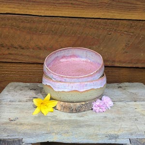 Könnte beinhalten: Eine runde Keramikschale mit gesprenkelter hellgrüner Basis und glattem, glänzendem rosa Innenraum. Die Glasur läuft an den Seiten herab und erzeugt einen Schichteffekt. Die Schale steht auf einer Holzoberfläche neben einer gelben und einer kleinen lila Blume.