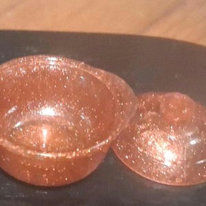 May include: A small, round, orange-colored container with a lid. The container and lid are covered in glitter. The container has a handle on one side. The lid is oval-shaped.