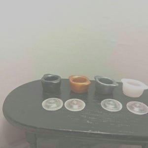 May include: A collection of miniature ceramic bowls in black, orange, gray, and white, with matching clear lids. The bowls are arranged on a dark oval table, showcasing a variety of colors and textures.