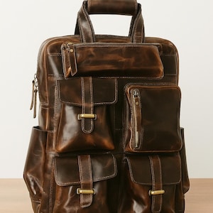 May include: A brown leather backpack with multiple pockets and compartments. The bag features a top handle, adjustable straps, and several exterior pockets with metal clasps and zips. The leather has a distressed finish.