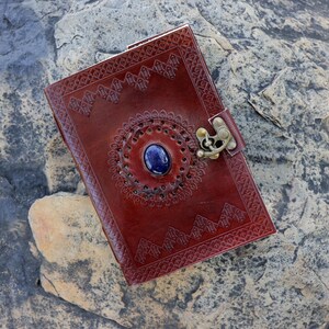 May include: A handcrafted, reddish-brown leather journal with intricate embossed patterns and a central blue gemstone. The journal features a decorative metal clasp and is closed. The cover is adorned with detailed geometric designs.