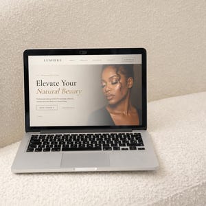 May include: A silver laptop displaying a website for Lumiere with the text Elevate Your Natural Beauty and a photograph of a person. The screen features navigation menus, a booking button, and professional makeup artist service details.