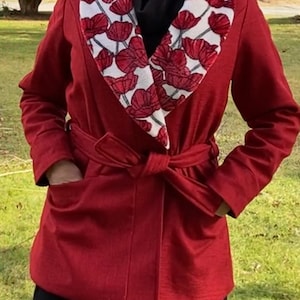 May include: A red robe with a white collar featuring a red floral pattern. The robe has a matching belt tied at the waist and pockets. The person is wearing a black top underneath.