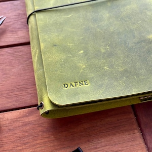 K&ouml;nnte beinhalten: Ein gr&uuml;nes Lederjournal mit dem Namen "DAFNE" auf dem Einband. Das Journal wird mit einem schwarzen Gummiband verschlossen. Zwei schwarze B&uuml;roklammern und eine silberne Schere sind ebenfalls zu sehen.