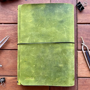 K&ouml;nnte beinhalten: Ein gr&uuml;nes Lederjournal mit dem Namen "DAFNE" auf dem Einband. Das Journal wird mit einem schwarzen Gummiband verschlossen. Verschiedene Bastelwerkzeuge und B&uuml;roklammern sind um das Journal herum auf einer Holzoberfl&auml;che angeordnet.