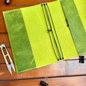 May include: A lime green leather notebook cover featuring a textured surface, light green stitching along the edges, and three black elastic cords threaded through metal eyelets for securing inserts, presented in an open, flat orientation.