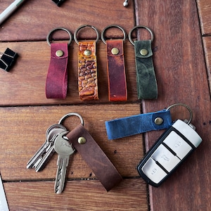 May include: A collection of leather keychains in various colors, including burgundy, brown, and blue, with metal key rings and rivets. Some keychains have textured leather. Keys and a car key fob are also present.